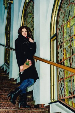 Kışlık elbiseli güzel kızıl saçlı bir kız elinde bir buket gülle bir binanın merdivenlerinde duruyor ve gülümsüyor. Kış duygusal portresi.