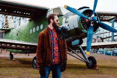 Eski bir uçağın arka planında sakallı genç bir pilot..