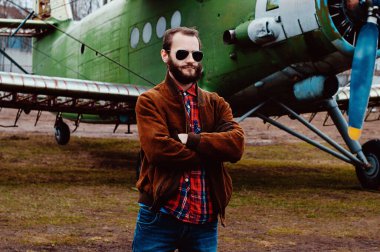 Eski bir uçağın arka planında sakallı genç bir pilot..