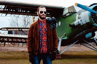 Eski bir uçağın arka planında sakallı genç bir pilot..