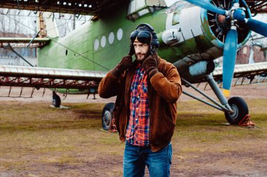 Eski bir uçağın arka planında sakallı genç bir pilot..