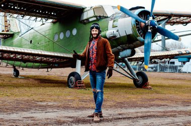 Eski bir uçağın arka planında sakallı genç bir pilot..