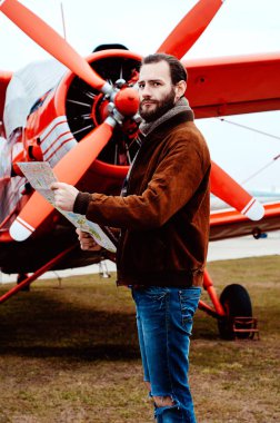 Sakallı genç bir pilot, bir gezgin kırmızı eski moda bir uçağın arka planında duruyor ve harita üzerinde çalışıyor..