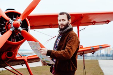 Sakallı genç bir pilot, bir gezgin kırmızı eski moda bir uçağın arka planında duruyor ve harita üzerinde çalışıyor..