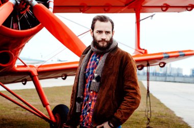 Sakallı genç bir pilot, bir gezgin kırmızı bir uçağın arka planında duruyor..