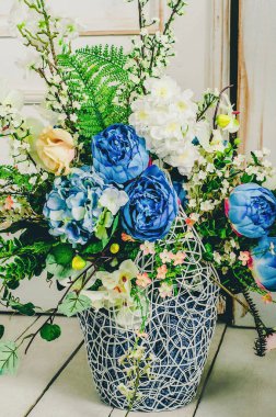 Beyaz orkidelerin, mavi güllerin ve eğreltiotu dallarının yanında dekoratif beyaz bir şöminenin üzerinde klasik bir kafes..