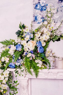 Beyaz orkidelerin, mavi güllerin ve eğreltiotu dallarının yanında dekoratif beyaz bir şöminenin üzerinde klasik bir kafes..