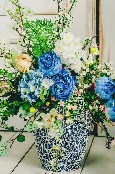 Beyaz orkidelerin, mavi güllerin ve eğreltiotu dallarının yanında dekoratif beyaz bir şöminenin üzerinde klasik bir kafes..