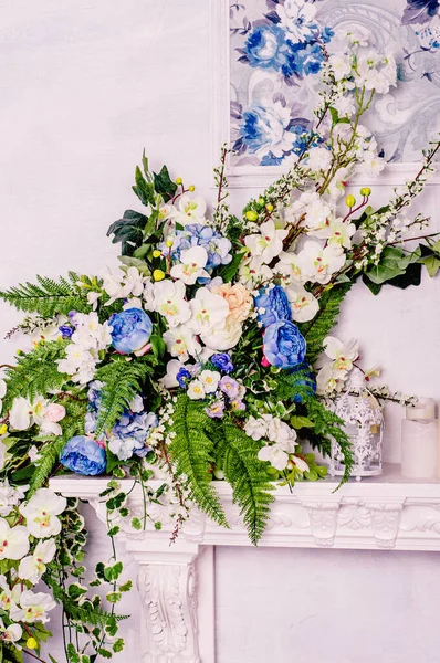 Beyaz orkidelerin, mavi güllerin ve eğreltiotu dallarının yanında dekoratif beyaz bir şöminenin üzerinde klasik bir kafes..