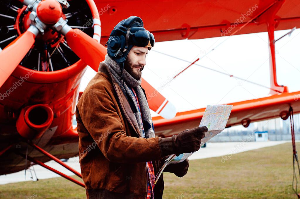 A bearded young pilot, a traveler stands on the background of a red old-fashioned airplane and studies the map.
