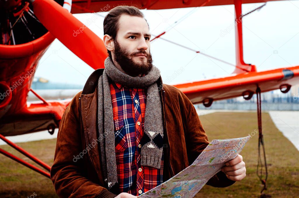 A bearded young pilot, a traveler stands on the background of a red old-fashioned airplane and studies the map.