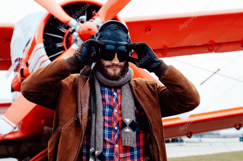 A bearded young pilot, a traveler stands on the background of a red vintage aircraft.