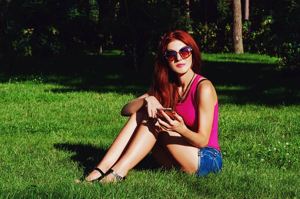公園の芝生の上には赤い長い髪の美しい女の子が座っており 彼女は彼女の手に電話を持っています 感情的な写真 — ストック写真