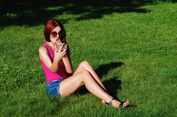 公園の芝生の上には赤い長い髪の美しい女の子が座っており 彼女は彼女の手に電話を持っています 感情的な写真 — ストック写真