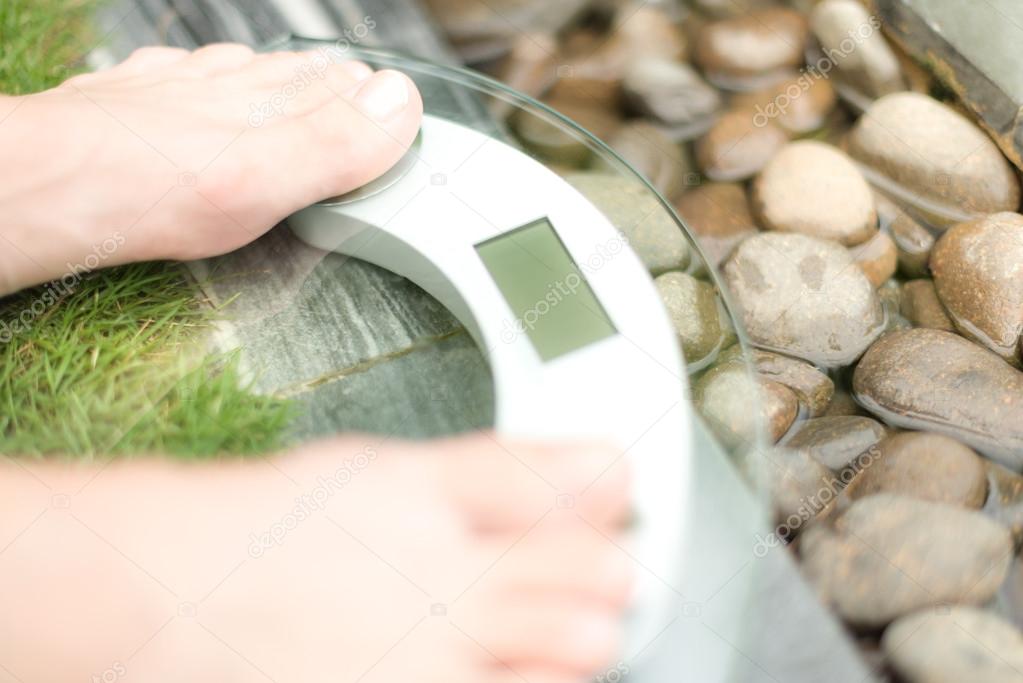 Feet on scale over green lawn and wet pebble.