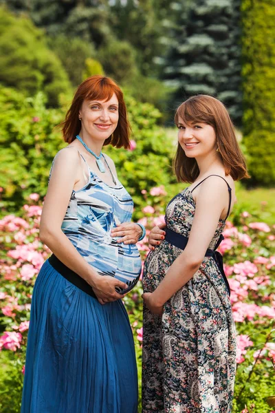 Zwei schöne lächelnde schwangere Freundinnen. Draußen. vor dem Hintergrund der Blumen — Stockfoto