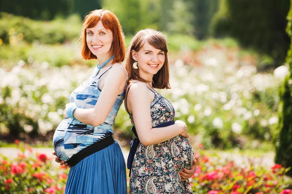 Две красивые молодые улыбающиеся беременные женщины. на улице. на фоне цветов — стоковое фото