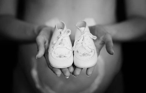 Mulher grávida segurando pequenos sapatos de bebê na frente da barriga — Fotografia de Stock