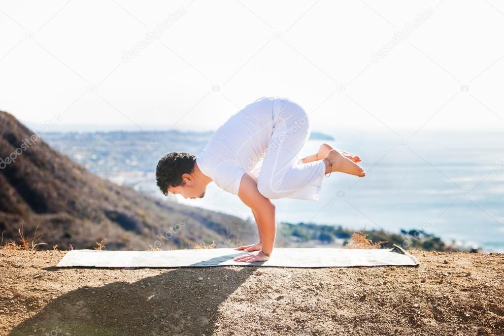 Asian man meditates in yoga position on high mountains above blue sky.