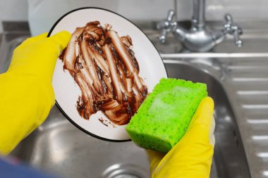 Eldiven giymiş insan elleri lavabodaki musluğun altında kirli bir tabağı süngerle yıkıyor..