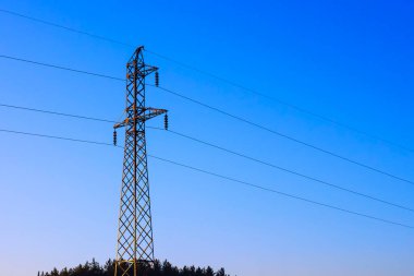 A metal power line tower reaches high into the sky, silhouetted against a clear blue background. Trees line the horizon, creating a peaceful rural setting at dusk.