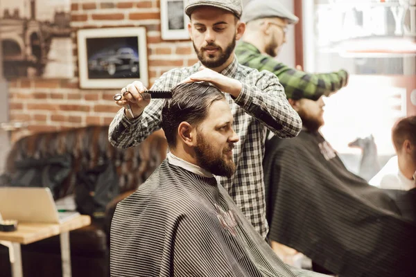Corte de cabelo na barbearia. Homem barbudo jovem recebendo corte de cabelo enquanto sentado na cadeira na barbearia. Cabeleireiro corta cabelo com tesoura e um pente — Fotografia de Stock