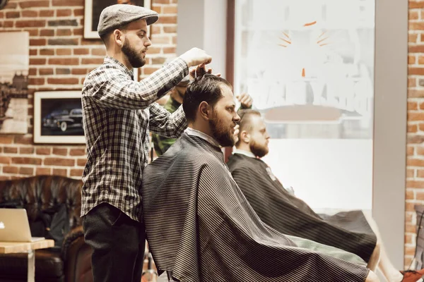 Corte de cabelo na barbearia. Homem barbudo jovem recebendo corte de cabelo enquanto sentado na cadeira na barbearia. Cabeleireiro corta cabelo com tesoura e um pente — Fotografia de Stock