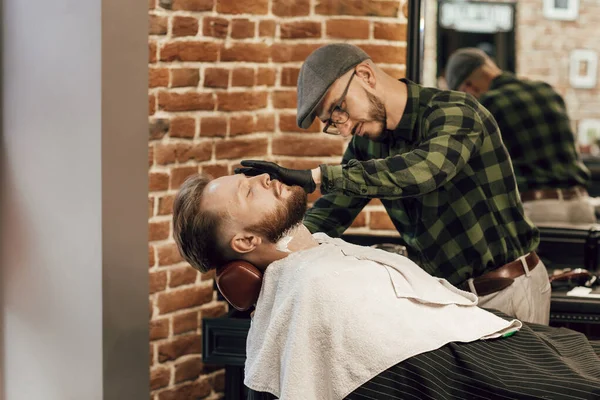 一个留着胡子的男人坐在理发店的扶手椅上，理发师给他刮胡子。理发店里的特写镜头，戴着手套的理发师用剃须刀刮胡子 — 图库照片