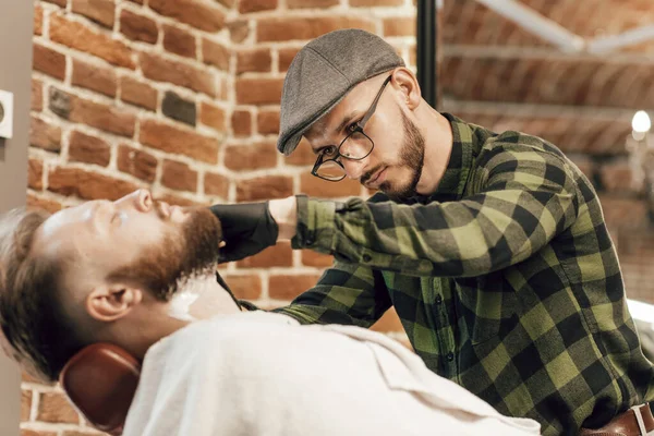 Homem barbudo sentado numa poltrona numa barbearia enquanto o cabeleireiro rapa a barba. O trabalho de close-up em uma barbearia, um cabeleireiro em luvas rapa a barba de clientes com uma navalha — Fotografia de Stock