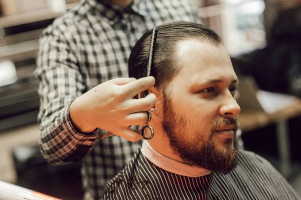 关闭理发店的理发 年轻的留着胡子的男人坐在理发店的椅子上理发 理发师用剪刀和梳子剪头发 — 图库照片