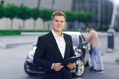 Takım elbiseli genç bir araba satıcısının yakın plan portresi. Arkasında genç bir çift yeni bir arabayı inceliyor..