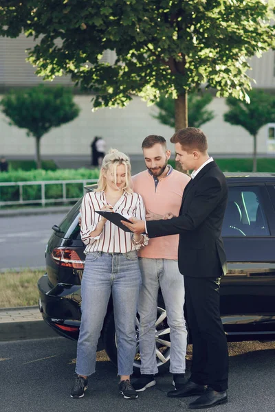 Seorang salesman muda menunjukkan mobil baru kepada pelanggan. Pasangan bahagia, pria dan wanita membeli mobil baru. Anak muda menandatangani dokumen untuk membeli mobil — Stok Foto