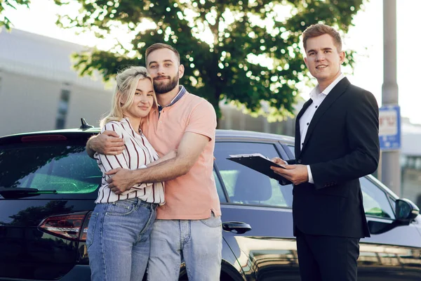 Salesman dan pasangan muda bahagia dengan mobil baru. Pasangan muda berdiri merangkul dekat mobil di luar. Membeli mobil — Stok Foto