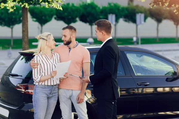 Młody Sprzedawca Pokazuje Klientom Nowy Samochód Szczęśliwa Para Mężczyzna Kobieta — Zdjęcie stockowe