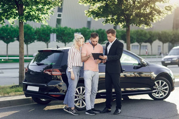 Un joven vendedor muestra un coche nuevo a los clientes. Pareja feliz, hombre y mujer compran un coche nuevo. Los jóvenes firman documentos para comprar un coche Imagen De Stock