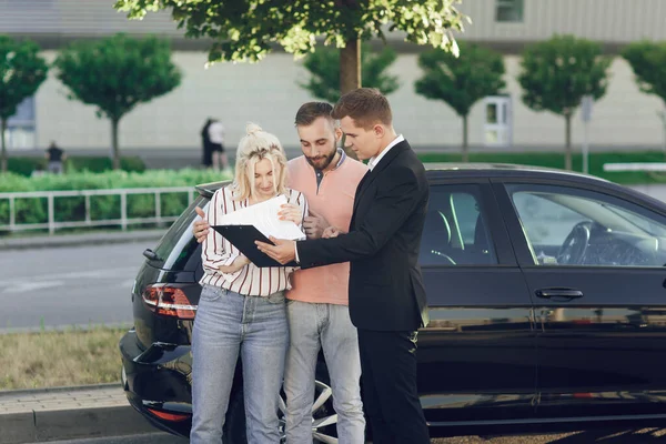 Un giovane venditore mostra una nuova auto ai clienti. Coppia felice, uomo e donna comprano una macchina nuova. I giovani firmano documenti per acquistare un'auto Fotografia Stock