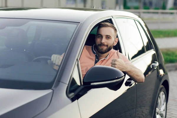 Primer plano del joven sonriendo y mostrando los pulgares hacia arriba en su coche nuevo. Comprar un coche Imágenes De Stock Sin Royalties Gratis