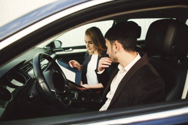 Genç bir adam araba kiralıyor. Satış merkezinin çalışanı arabada belgeler gösteriyor..