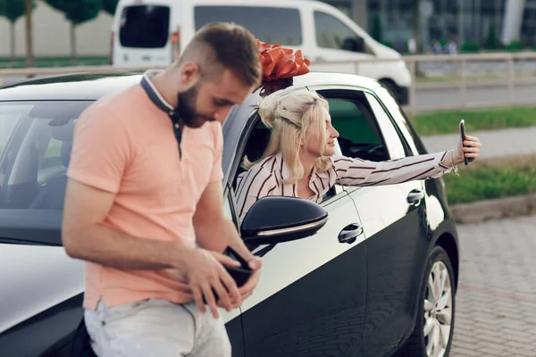 Mężczyzna daje kobiecie niespodziankę, kupując nowy samochód. Młoda szczęśliwa kobieta siedzi za kółkiem i robi sobie selfie — Zdjęcie stockowe