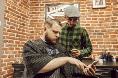 Berber dükkanında sakallı bir adam elinde cep telefonuyla kuaföre saç stilini gösteriyor. Berber dükkanındaki adamlar.