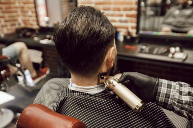 Genç sakallı adam berber dükkanında sandalyede otururken saçını kestiriyor. Kuaför sakalını kırpıcı ile keser.