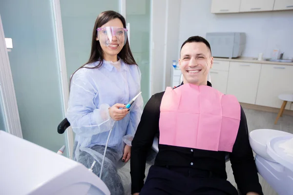 Un joven y una doctora en el consultorio de dentistas, sentados y sonriendo, mirando a la cámara. Mujer sosteniendo una herramienta de tratamiento dental —  Fotos de Stock