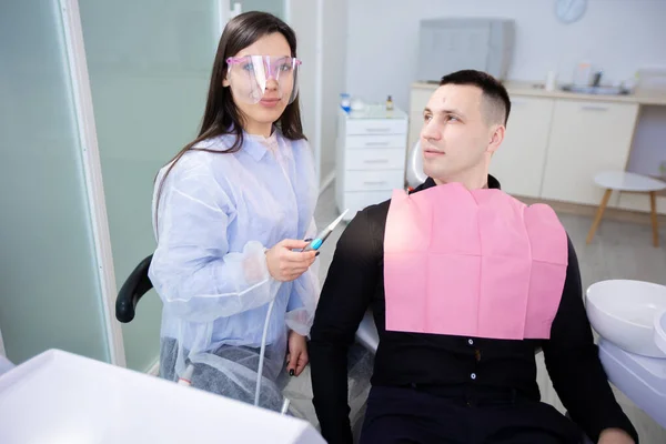 Young man and female doctor in dentists office, sitting and smiling, looking at camera. Woman holding a dental treatment tool — Stock Photo, Image