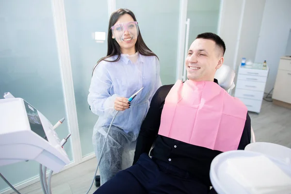 Giovane medico nello studio dentistico, seduto e sorridente, guardando la macchina fotografica. Donna in possesso di uno strumento di trattamento dentale — Foto Stock