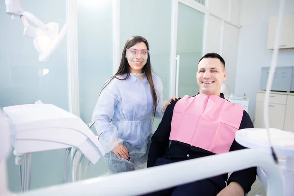 Dişçi muayenehanesinde oturan ve gülümseyen genç adam ve bayan doktor kameraya bakıyor. Kadının elinde diş tedavisi aracı var. — Stok fotoğraf