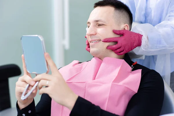 Smiling young man looking at mirror in the dentists chair. Man in the dental office after hygiene, prevention of dental diseases — Stock Photo, Image