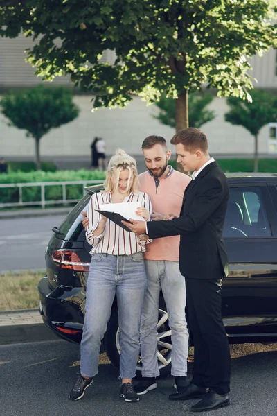 Seorang salesman muda menunjukkan mobil baru kepada pelanggan. Pasangan bahagia, pria dan wanita membeli mobil baru. Anak muda menandatangani dokumen untuk membeli mobil — Stok Foto