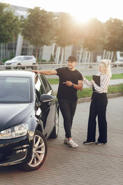 Młoda blond kobieta konsultant pokazuje klientowi samochód na zewnątrz. Mężczyzna kupuje samochód. Test jazdy nowy samochód — Zdjęcie stockowe