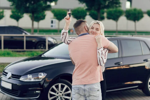 Muž dává překvapení ženě tím, že si koupí nové auto. Mladý pár si koupí auto, muž a žena se objímají poblíž auta na ulici. Žena držící klíčky od auta v rukou — Stock fotografie