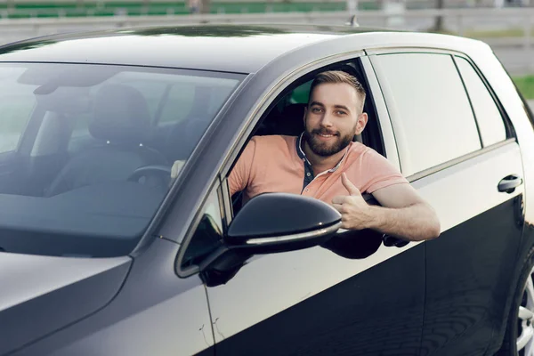 Zbliżenie młodzieńca uśmiechającego się i pokazującego kciuki w swoim nowym samochodzie. Kupowanie samochodu — Zdjęcie stockowe